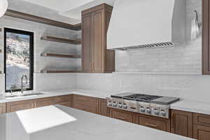 Kitchen featuring premium range hood, open shelves, stainless steel gas cooktop, light stone counters, and decorative backsplash