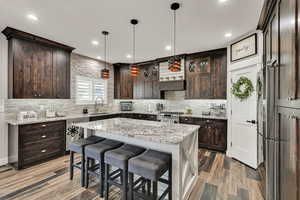 Kitchen featuring backsplash, dark brown cabinets, a kitchen island, wood finished floors, and decorative light fixtures