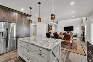 Kitchen featuring stainless steel fridge, dark brown cabinetry, light stone countertops, a kitchen island, and wood finished floors