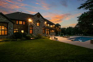 Back of property at dusk featuring stone siding, a lawn, an outdoor pool, and a patio