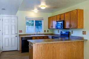 Kitchen with a textured ceiling, a peninsula, brown cabinetry, black appliances, and dark wood-type flooring