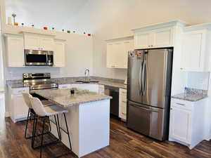 Kitchen featuring appliances with stainless steel finishes, light stone counters, white cabinetry, dark wood-type flooring, and a kitchen bar