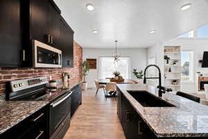 Kitchen with dark cabinets, appliances with stainless steel finishes, dark stone countertops, hanging light fixtures, and light wood-style flooring