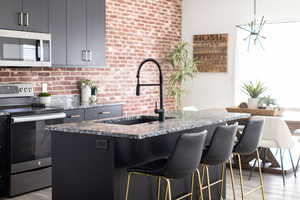 Kitchen featuring appliances with stainless steel finishes, light wood-type flooring, dark stone countertops, a kitchen island with sink, and brick wall