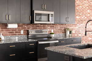 Kitchen featuring appliances with stainless steel finishes, light stone counters, modern cabinets, dark cabinetry, and brick wall