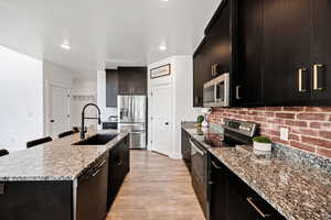 Kitchen with appliances with stainless steel finishes, light stone counters, light wood finished floors, a center island with sink, and dark cabinets