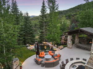 Patio terrace at dusk with a patio, an outdoor fire pit, a wooded view, and an outdoor stone fireplace
