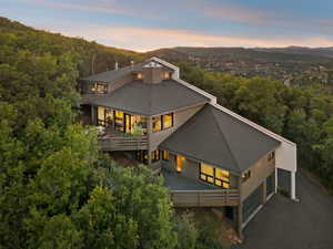 Rear view of property featuring a balcony, driveway, a view of trees, a shingled roof, and a patio area