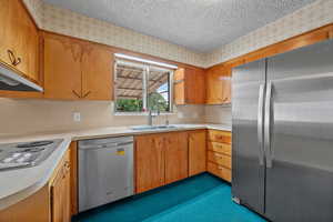 Kitchen with appliances with stainless steel finishes, light countertops, a textured ceiling, brown cabinetry, and under cabinet range hood