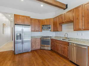 Kitchen with appliances with stainless steel finishes, light stone counters, brown cabinets, recessed lighting, and beam ceiling