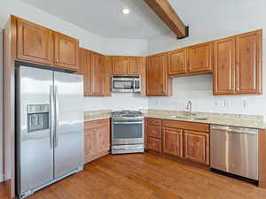 Kitchen with stainless steel appliances, brown cabinets, beamed ceiling, dark wood-style floors, and light stone counters