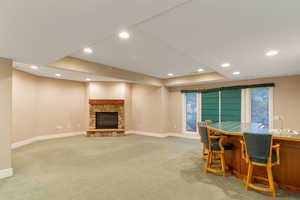 Dining space with a stone fireplace, light colored carpet, recessed lighting, bar with sink, and a tray ceiling