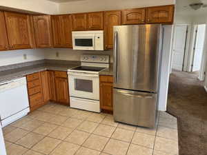 Kitchen featuring white appliances, brown cabinetry, light tile patterned floors, and light colored carpet
