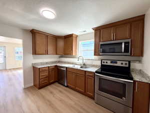 Kitchen featuring stainless steel appliances, brown cabinetry, a textured ceiling, light wood-type flooring, and light stone countertops