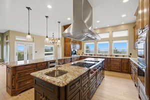 Kitchen featuring an island with sink, pendant lighting, light wood finished floors, light stone counters, and island range hood