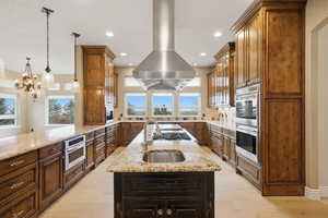 Kitchen featuring light stone counters, island exhaust hood, an island with sink, stainless steel double oven, and hanging light fixtures