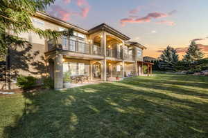 Back of house at dusk featuring a balcony, stucco siding, a patio area, and a lawn