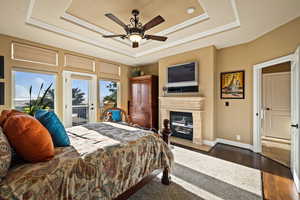 Bedroom with access to exterior, a tray ceiling, dark wood-style floors, a glass covered fireplace, and a ceiling fan