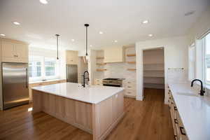Kitchen featuring open shelves, high end appliances, backsplash, light LVP floors, and an island with sink