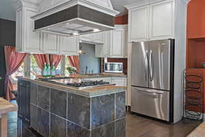 Kitchen with custom exhaust hood, appliances with stainless steel finishes, white cabinetry, dark wood-style flooring, and a textured ceiling