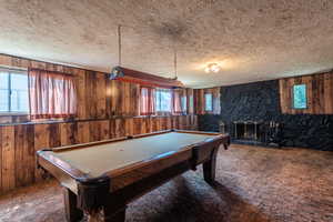 Extra large family room with daylight above ground windows, wood paneling walls, a pool table, a stone wood burning fireplace, carpet flooring, and a textured ceiling