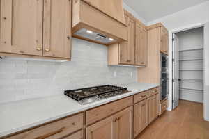 Kitchen with decorative backsplash, light brown cabinets, light wood-style flooring, premium range hood, and appliances with stainless steel finishes