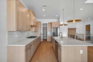 Kitchen with light brown cabinetry, decorative backsplash, light wood-style floors, a center island with sink, and light stone countertops
