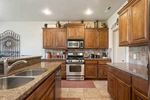 Kitchen featuring brown cabinetry, stainless steel appliances, tasteful backsplash, dark stone counters, and light tile patterned floors