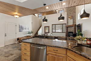 Kitchen with wooden ceiling, dark stone countertops, dishwasher, open floor plan, and a stone fireplace