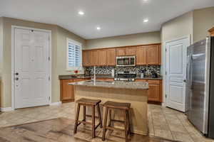 Kitchen featuring stainless steel appliances, backsplash, light stone counters, light tile patterned floors, and brown cabinets