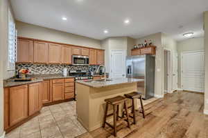 Kitchen featuring backsplash, appliances with stainless steel finishes, a kitchen breakfast bar, light stone countertops, and an island with sink