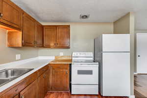 Kitchen featuring white appliances, light countertops, a textured ceiling, brown cabinetry, and light wood-style floors