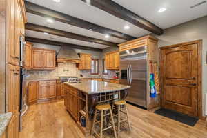 Kitchen with tasteful backsplash, light wood-style flooring, appliances with stainless steel finishes, beamed ceiling, and an island with sink