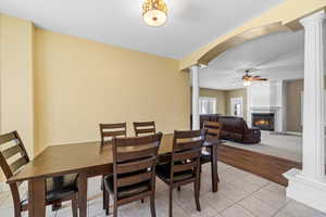 Dining room featuring ornate columns, light tile patterned floors, ceiling fan, a large fireplace, and arched walkways