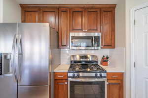 Kitchen featuring appliances with stainless steel finishes, brown cabinetry, decorative backsplash, and light quartz countertops