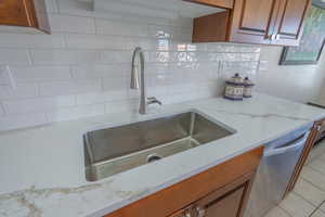 Kitchen with brown cabinetry, decorative backsplash, light quartz countertops, stainless steel sink, dishwasher, and light tile patterned floors