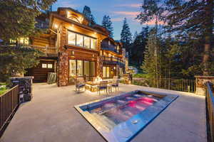 Back of property at dusk featuring a patio, a fire pit, a chimney, an in-ground hot tub, and stone siding