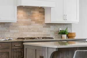 Kitchen with decorative backsplash, custom exhaust hood, white cabinetry, stainless steel gas stovetop, and light stone counters