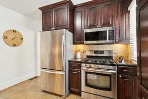 Kitchen featuring stainless steel appliances, tasteful backsplash, light tile patterned floors, dark stone counters, and dark brown cabinets