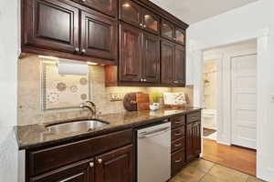 Kitchen with dark brown cabinets, decorative backsplash, dark stone countertops, stainless steel dishwasher, and light tile patterned floors