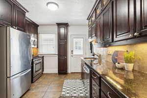 Kitchen with dark brown cabinets, stainless steel appliances, dark stone countertops, a textured ceiling, and decorative backsplash