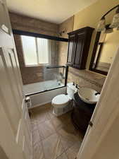 Full bathroom with bath / shower combo with glass door, vanity, light tile patterned floors, a textured ceiling, and tile walls