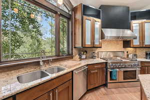 Kitchen featuring decorative backsplash, appliances with stainless steel finishes, light stone counters, wall chimney range hood, and glass insert cabinets
