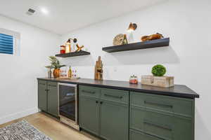 Indoor dry bar with green cabinetry, light wood-style flooring, wine cooler, open shelves, and recessed lighting
