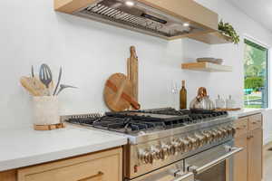Kitchen featuring custom exhaust hood, light brown cabinetry, light stone countertops, range with two ovens, and open shelves