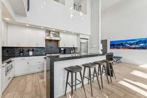 Kitchen with wood finish floors, a towering ceiling, tasteful backsplash, a kitchen bar, and wall chimney range hood