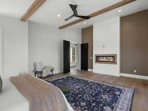 Bedroom featuring beam ceiling, a glass covered fireplace, wood finished floors, recessed lighting, and ceiling fan
