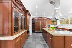 Kitchen with light stone counters, island range hood, an island with sink, dark tile patterned flooring, and brown cabinetry