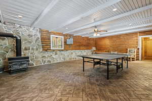 Rec room featuring a wood stove, a ceiling fan, beamed ceiling, rustic walls, and recessed lighting
