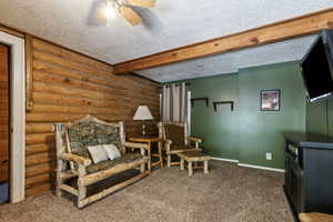 Living area featuring beamed ceiling, rustic walls, a textured ceiling, carpet floors, and ceiling fan